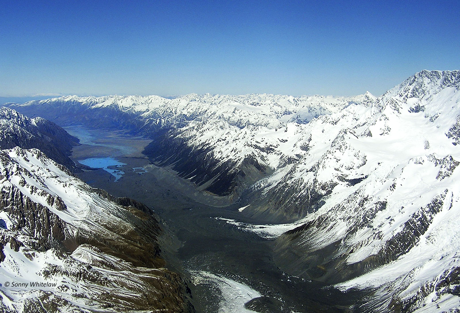 Facing South: Tasman Glacier (foreground – darker areas are moraine gravels over the ice) on the eastern side of Aoraki Mt Cook (right), and Tasman River (centre back).
